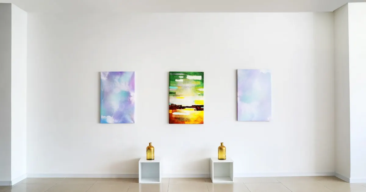 Three abstract canvases hung on a bare white gallery wall above two gold bottles on white plinths