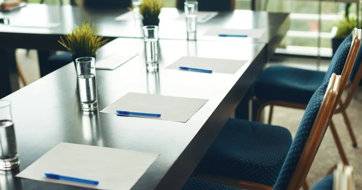 A long boardroom table set with blank notepads, blue pens and water glasses beside empty chairs