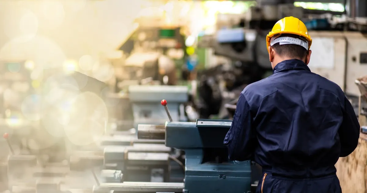 A worker in a yellow hard hat and blue overalls seen from behind, operating a row of lathes