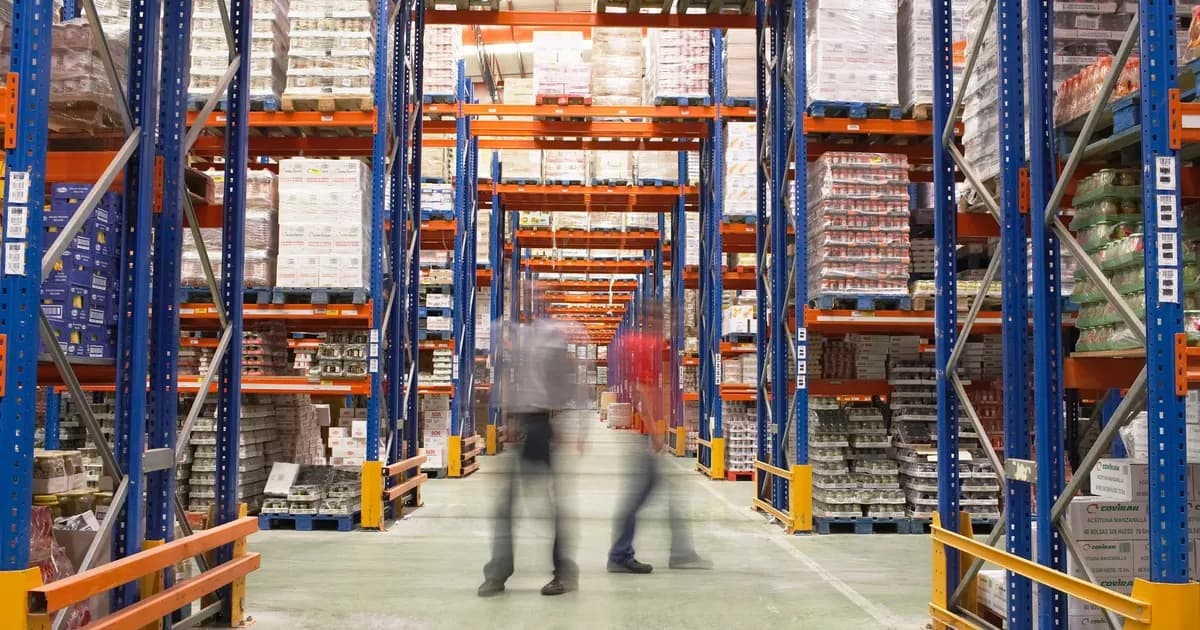 Two blurred figures walking down a warehouse aisle between tall racks of palletised goods