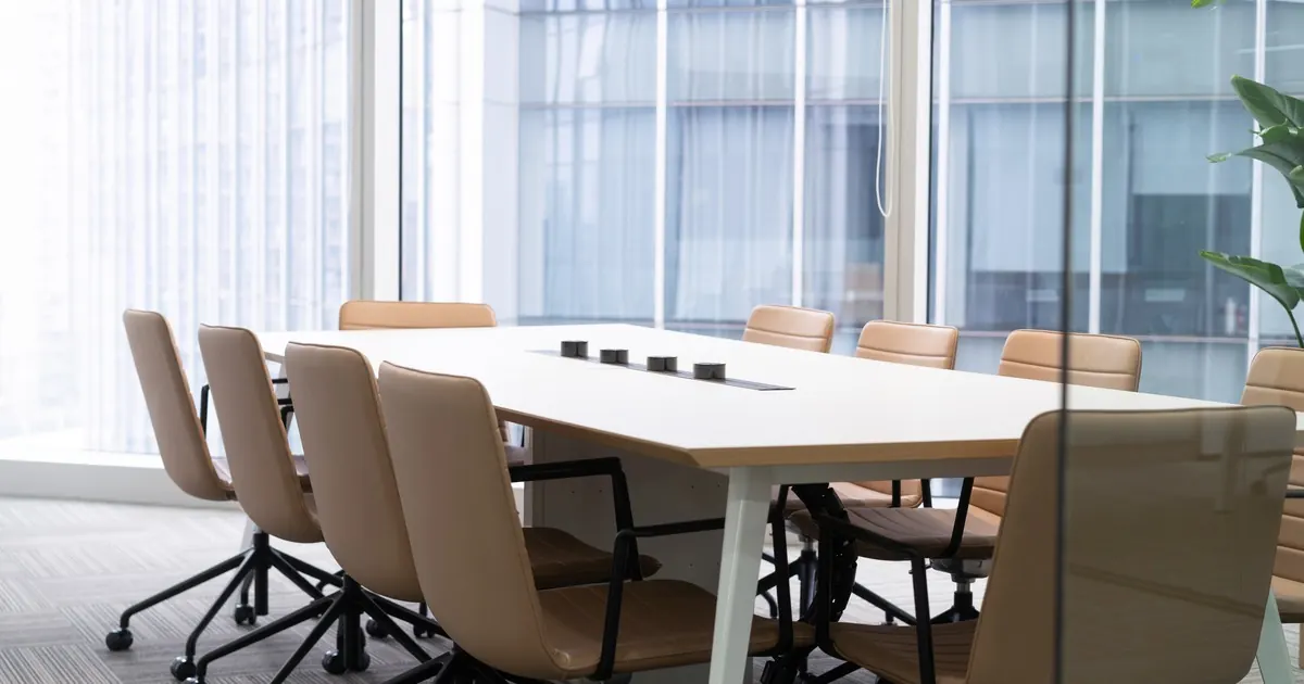 An empty meeting room with a long white table, tan leather chairs and floor-to-ceiling windows overlooking office towers