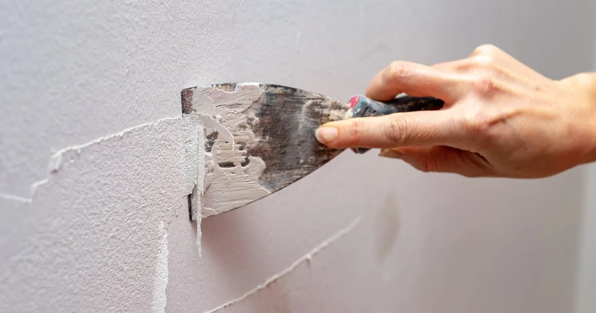 A hand scraping cracked paint and filler from a wall with a putty knife