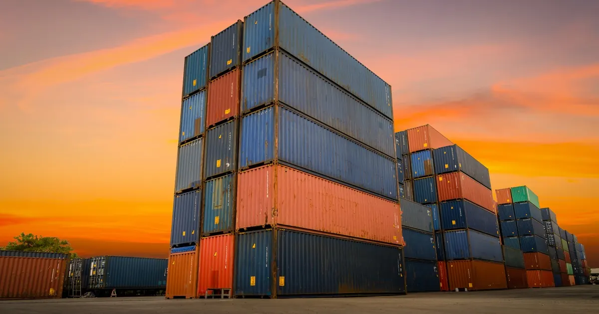 Stacks of shipping containers at a port terminal under an orange sunset sky