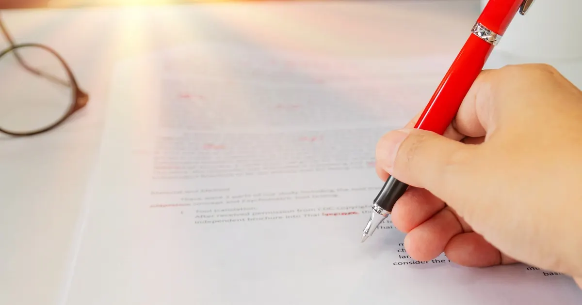 A hand holding a red pen above a printed document covered in red corrections, glasses alongside