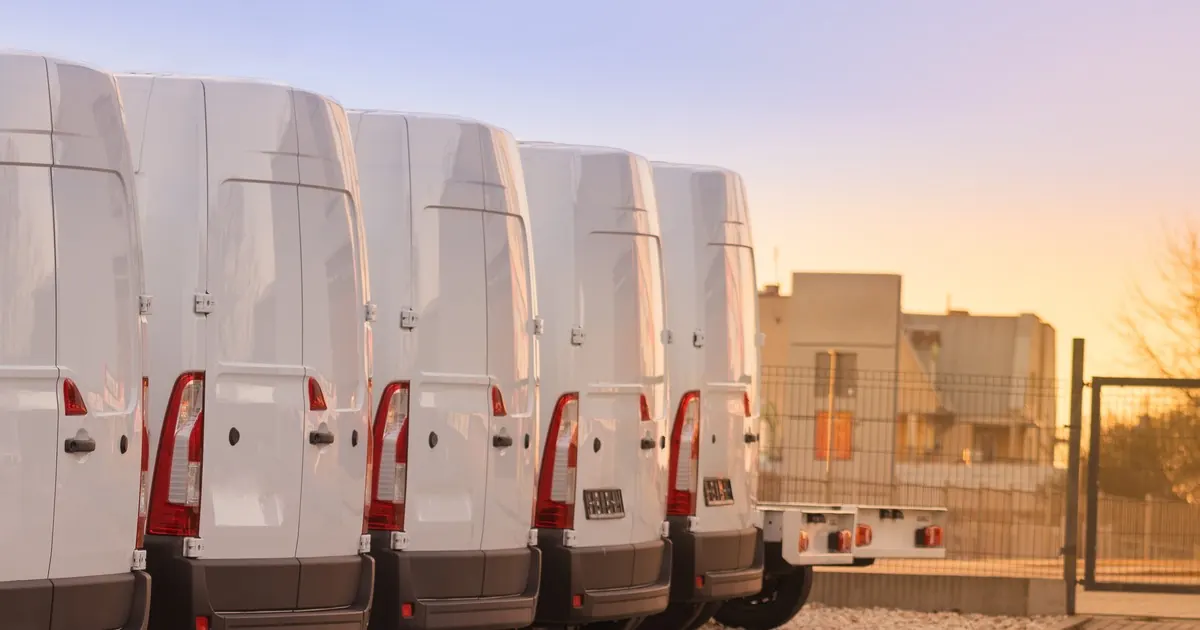 A row of plain white delivery vans parked rear-first in a fenced yard at dusk